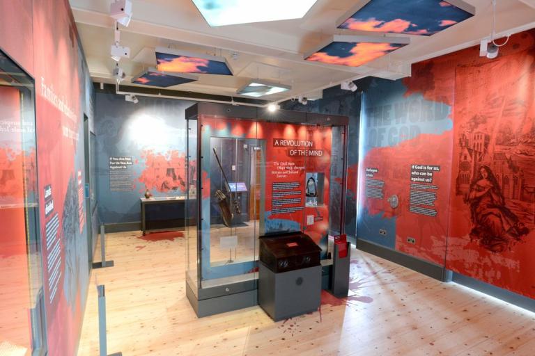 A gallery room showing red and black wallpaper that depicts information on historial conflict and creativity.